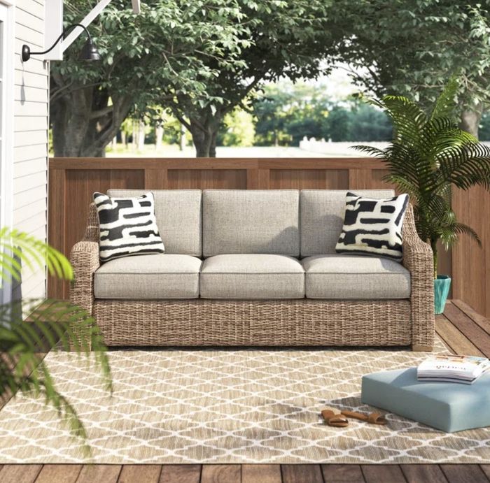 A warm and inviting outdoor patio showcasing a wicker sofa with two decorative throw pillows, a geometric-patterned rug, a vibrant blue ottoman, and lush plants, all shaded by the surrounding trees.