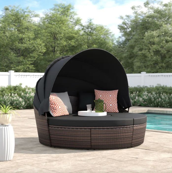 A dark wicker daybed with brown cushions and decorative pillows, draped with a matching dark brown cover, resting on a pool deck.