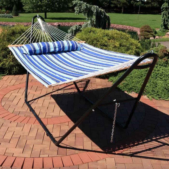 A blue striped hammock suspended on a black metal frame, resting on a brick patio surrounded by a lush garden.