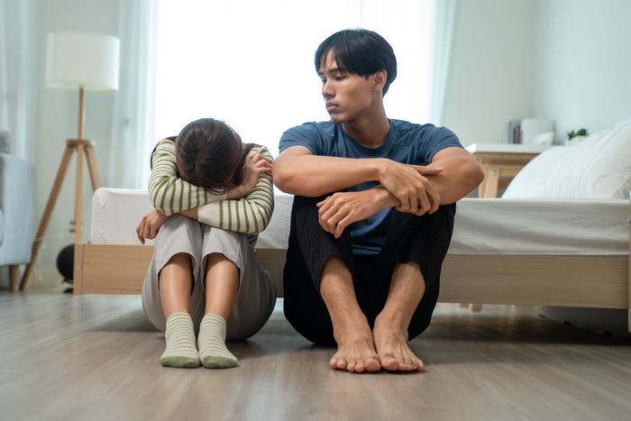 A couple sits on the floor by the bed, looking upset. One person rests their head on their knees while the other looks concerned.