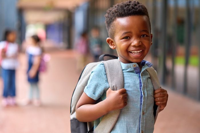 A child grinning, wearing a backpack, standing outdoors among other kids, with a blurred background