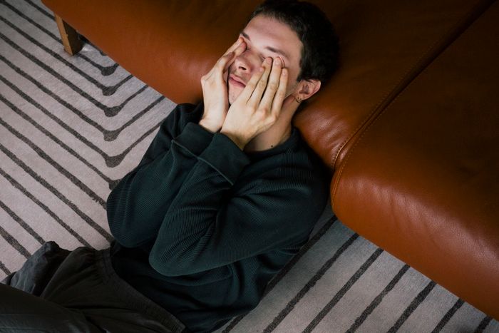 A person sitting on the floor, leaning against a couch, eyes closed, with their hands on their face, looking deep in thought or overwhelmed