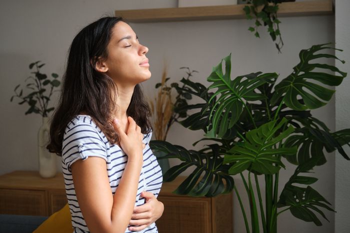 A woman in a striped shirt calmly breathes with her eyes shut, hand placed on her chest, surrounded by houseplants.
