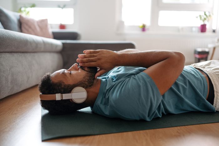 A person lying on a yoga mat with headphones on, looking relaxed, with their hands gently resting on their face, in a comfortable living room environment