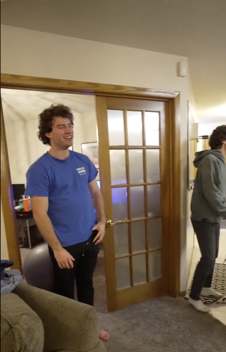 Two people standing indoors, smiling, in a casual environment with a slightly open door in the background