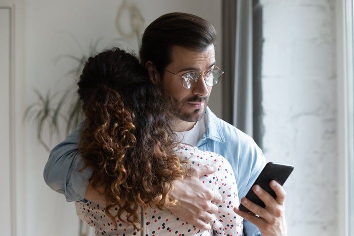 A man with glasses embraces a woman, both gazing at his phone with concerned expressions