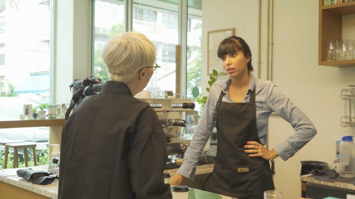 Two people in a coffee shop: one with gray hair facing the counter, the other with dark hair and an apron, standing behind the counter, having a conversation