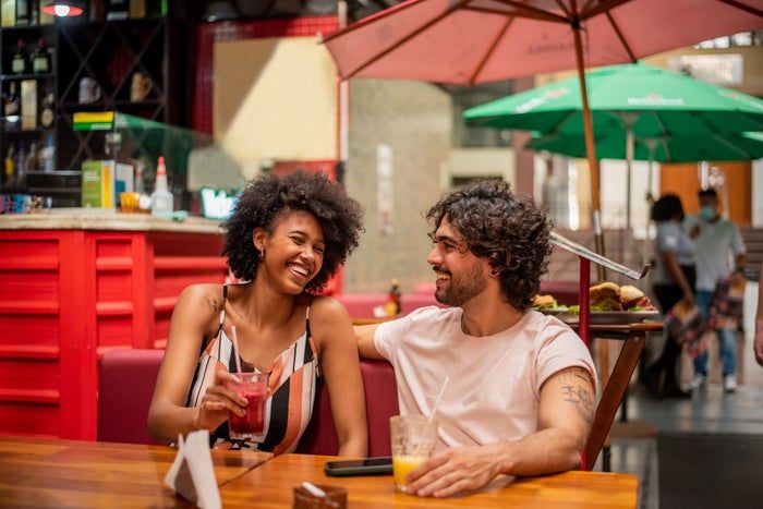 A woman and a man exchanging smiles while seated at an outdoor café table, each holding a drink
