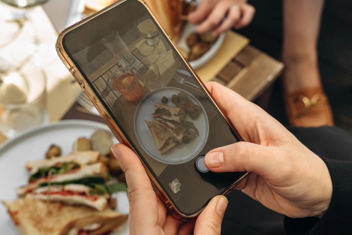 A person using a smartphone to photograph a table filled with various deli sandwiches in a casual dining setting.