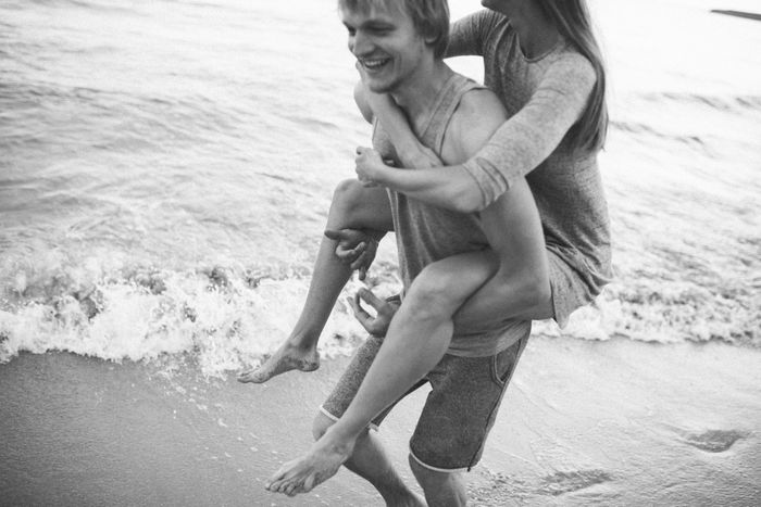 A man giving a woman a piggyback ride on the beach, both smiling, waves in the background