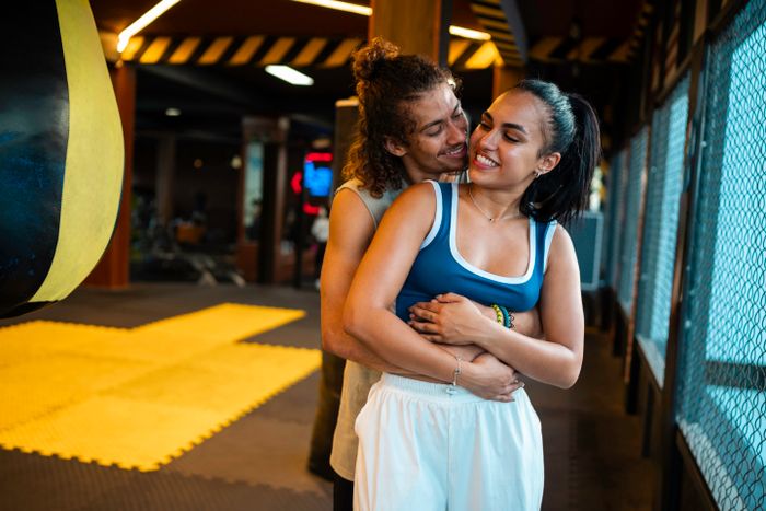 A couple in a gym shares a playful moment near a punching bag, the woman grinning while the man stands behind her, both dressed in casual workout clothes.