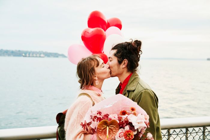 A couple shares a kiss near the water, surrounded by heart-shaped balloons and holding a bouquet of flowers.