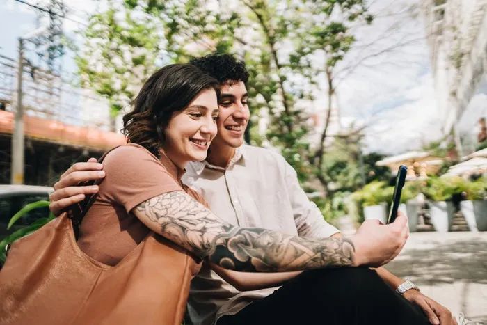 A couple sitting on a park bench, happily taking a selfie together. One partner has a tattoo on their arm, and they both appear to be enjoying a sunny day.