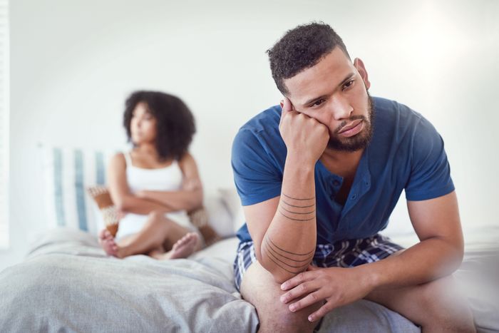 A man and a woman sitting apart on a bed, facing away from each other, looking upset
