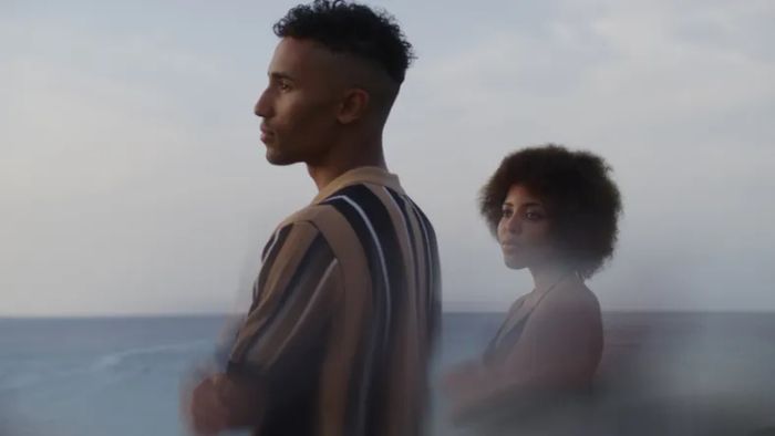 A man and woman stand together by the ocean, gazing thoughtfully into the distance.