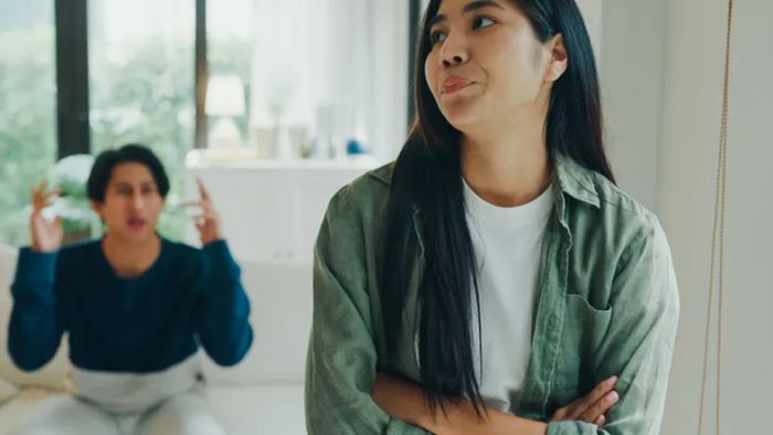 Two people at home; one looks away with arms crossed, while the other gestures in the background. The tension in the air is palpable.