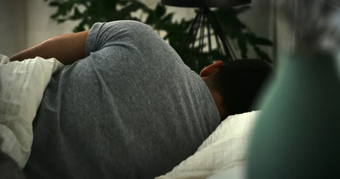 A person sleeping on their side in bed, partially covered by a blanket, with the room dimly lit.