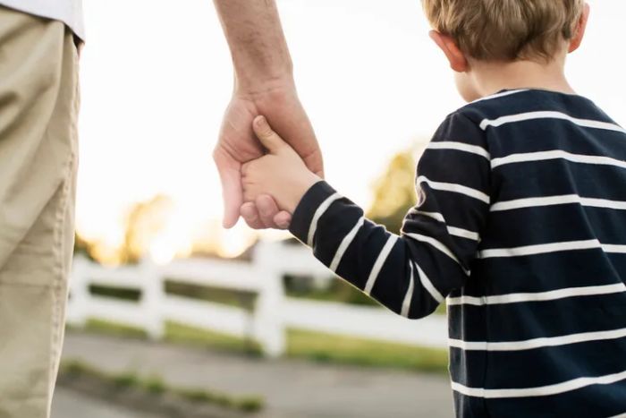 An adult and child walking hand-in-hand, with the child wearing a striped shirt. The background features a white fence and a blurred outdoor scene.