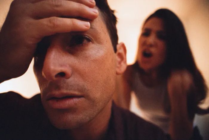 A person with their head in their hands, overwhelmed, while someone else shouts in the background.