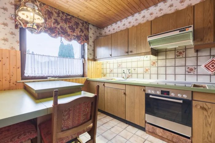 A retro-style kitchen featuring wooden cabinets, decorative tiles, a stove, and a table with chairs positioned beneath a window adorned with floral curtains.