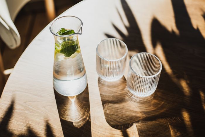 A glass pitcher filled with mint-infused water sits beside two empty glasses on a sunlit wooden table, evoking a refreshing vibe