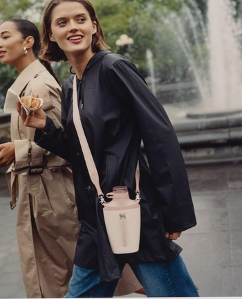 Two individuals stroll through an urban area; one of them is holding a food cone and sporting a bag, casual coats, and jeans.