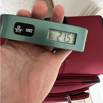 A hand holding a luggage scale with a red suitcase, showing a digital reading of 2.15 on the screen.