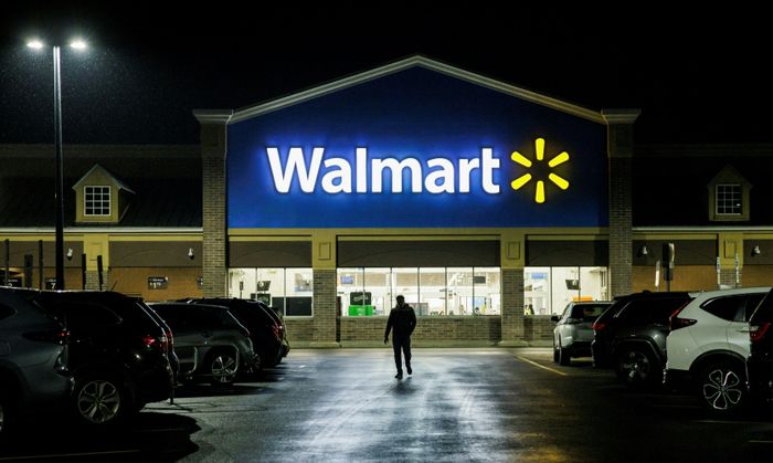 A person heads toward a Walmart store in the evening, passing rows of parked cars on either side.