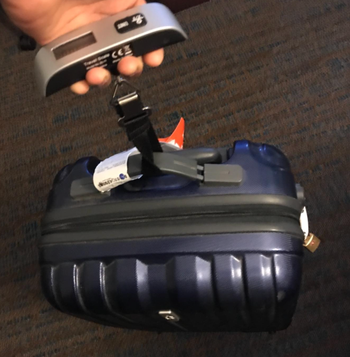 A hand holding a luggage scale, with a small black wheeled suitcase attached for weighing.