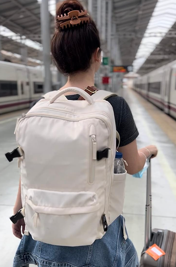 A reviewer carrying a white backpack with a front pocket and a side sleeve for a water bottle in an airport.