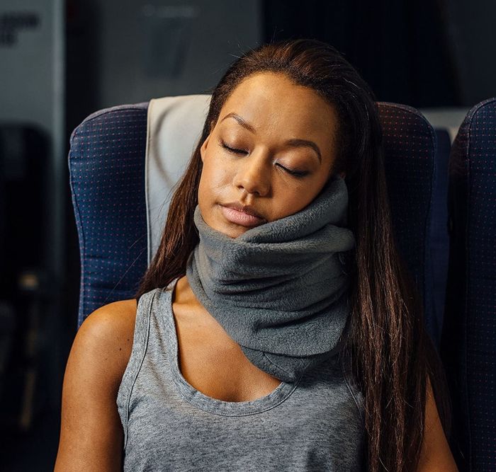 A model wearing a gray Trtl pillow while sleeping on a plane.