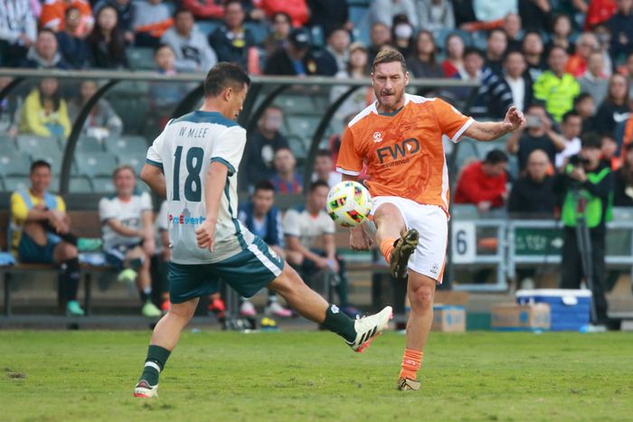 Two soccer players battle it out on the field: one in an orange jersey mid-kick, while the other in a gray jersey tries to block. Spectators cheer from the stands.