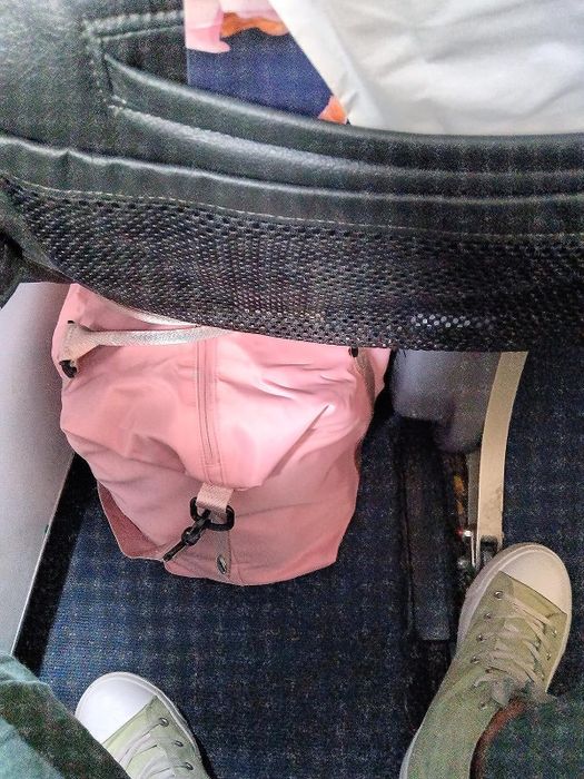 A reviewer showing off how perfectly their pink bag fits under the seat in front of them on the plane.