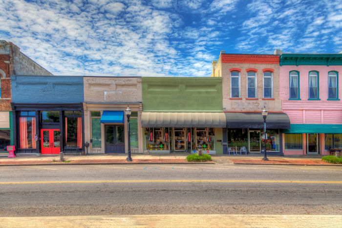 A peaceful street lined with charming, colorful storefronts, photographed under a bright sky dotted with fluffy clouds