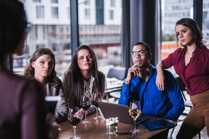 Four individuals in a contemporary office setting focus intently on a presenter; wine glasses on the table indicate a relaxed meeting atmosphere