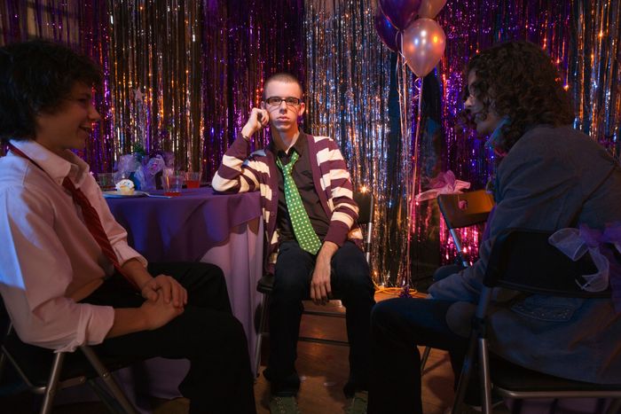 Three individuals sit at a table in a festive setting with glittering decorations and balloons, dressed casually in jackets and ties, engaged in lively conversation