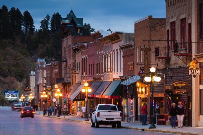 A charming street scene featuring historic shops, pedestrians strolling, and a mix of parked and moving vehicles