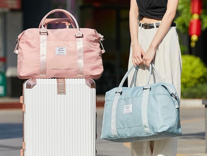 A model holding the blue duffel bag with the pink one resting on a suitcase beside them.