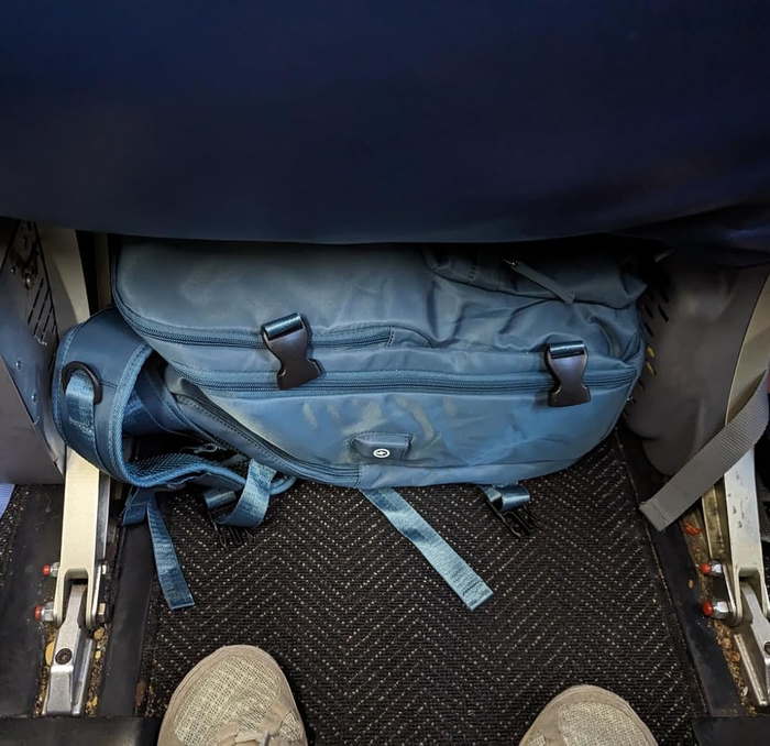 A reviewer demonstrating how their blue backpack fits perfectly under the airplane seat in front of them.