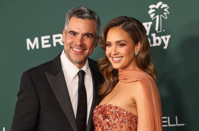 At a star-studded event, a man dressed in a suit and a woman wearing a stunning gown with a sheer cape pose on a green carpet.