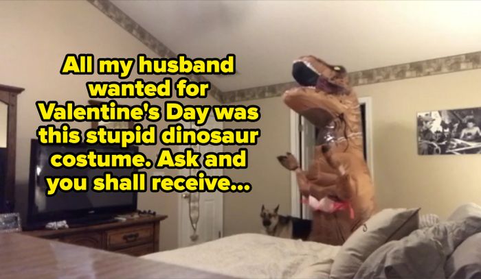 A person dressed in a dinosaur costume stands in a bedroom with a dog nearby.