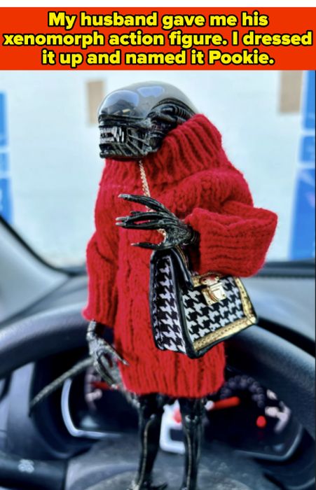 A xenomorph figurine dressed in a red knit sweater, carrying a houndstooth bag, and perched on a car dashboard