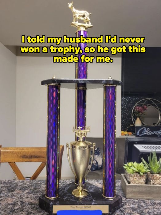 A tall trophy featuring a gold goat figure on top, purple columns, and a base inscribed with 'The Throat GOAT,' proudly displayed on a kitchen counter