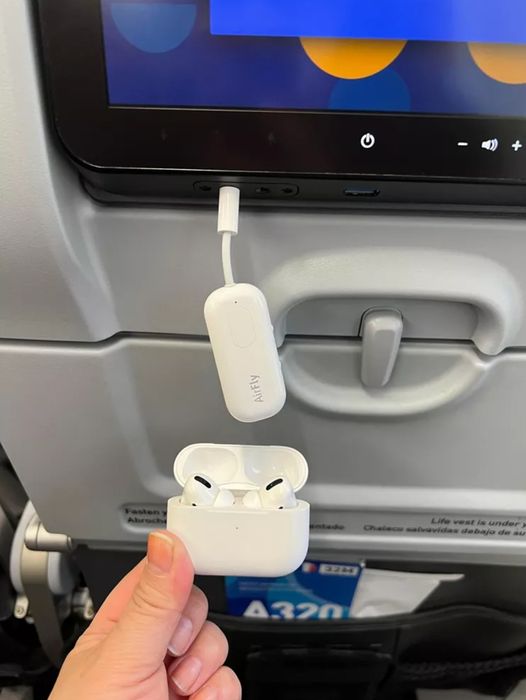 A person holding wireless earbuds below an airplane seat monitor, with an audio adapter plugged into the screen