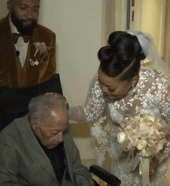 A bride adorned in a floral lace wedding gown gently caresses the head of an elderly man seated before her, while a man in a velvet suit stands close by.