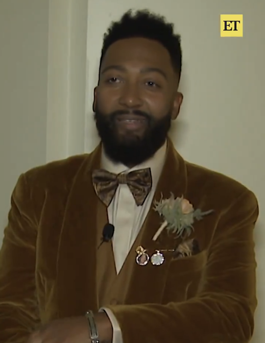 A man dressed in a velvet suit, complete with a bow tie and boutonniere, strikes a pose against a media backdrop.
