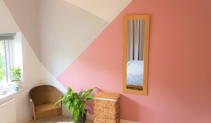 A modern, bright room featuring a wicker chair, a potted plant, a wicker basket, and a full-length mirror set against a wall with a geometric design.