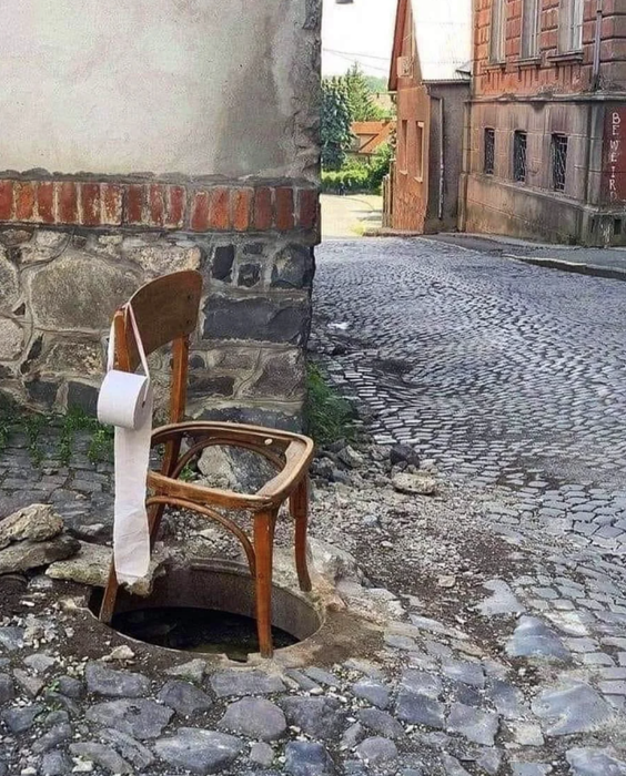 A makeshift toilet consisting of a chair with a hole, placed over an open manhole, complete with toilet paper attached