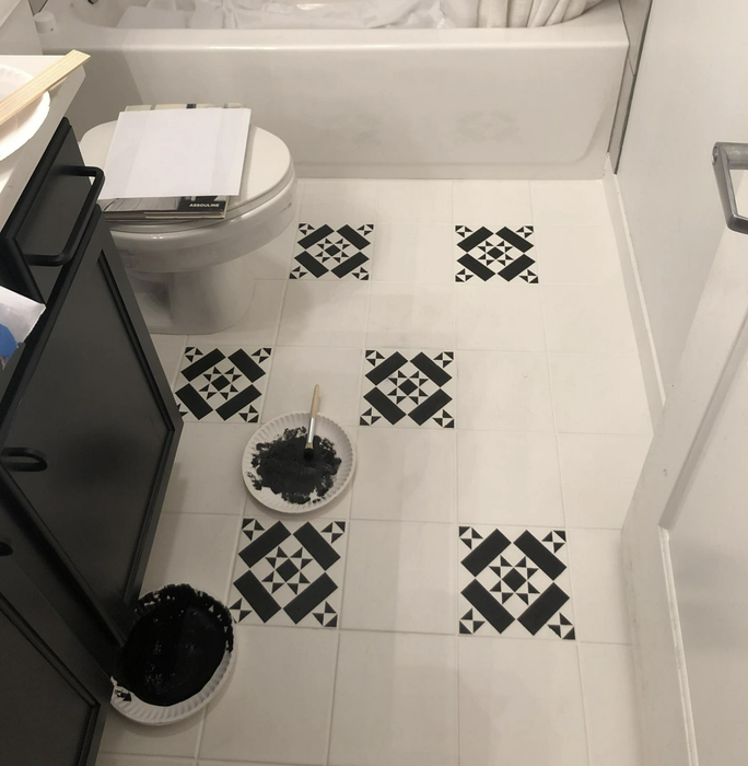A bathroom floor with someone performing a paint touch-up next to a toilet and trash bin