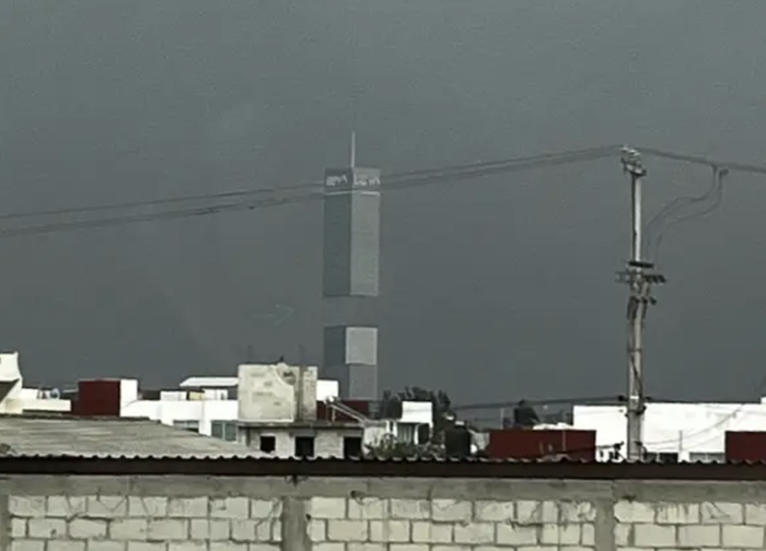 A towering skyscraper under construction in an urban setting, with scaffolding clearly visible. The foreground features rooftops and utility poles.